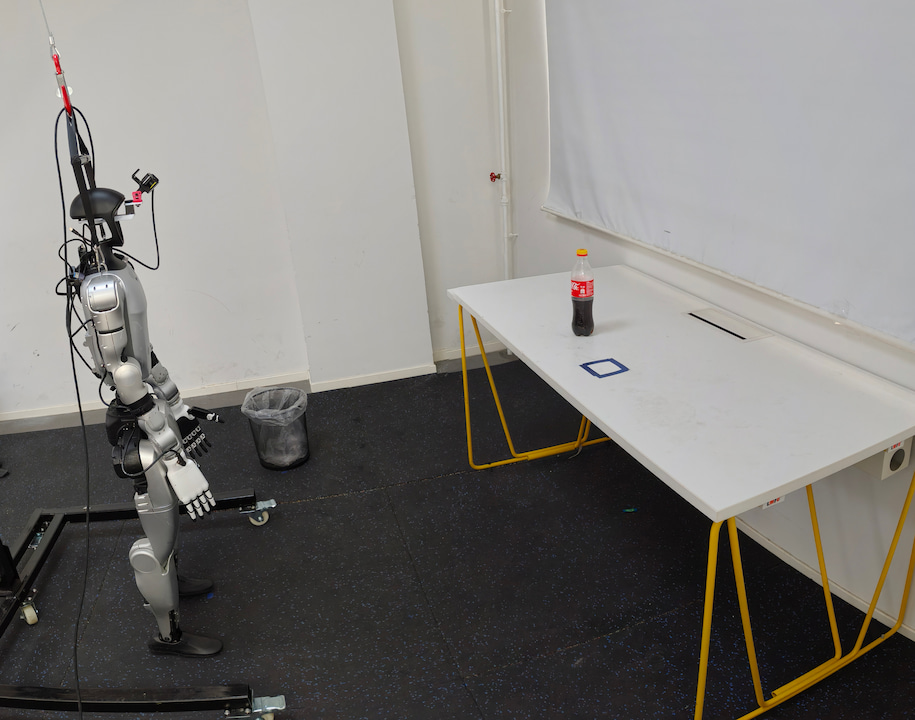 The Unitree G1 humanoid next to a table with a cola bottle and a nearby bin in the real-world experiment.